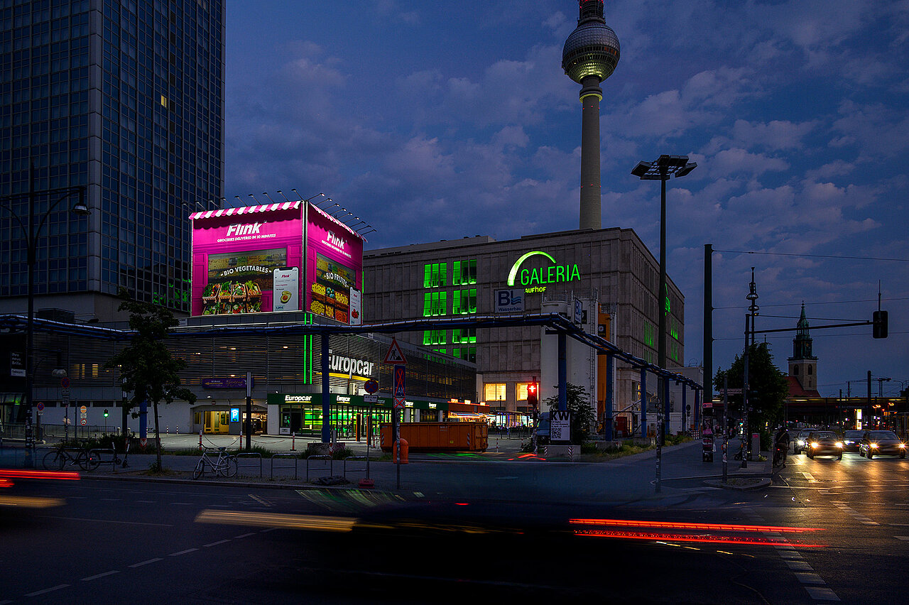 Nächtliche Stadtaufnahme am Berliner Alexanderplatz mit beleuchtetem pinken Flink-Werbekiosk auf einem Dach, daneben der Fernsehturm und die lichtdurchflutete Galeria.