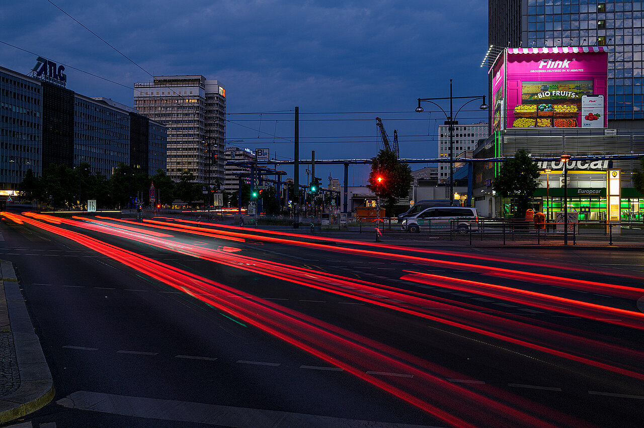 Nachtaufnahme am Berliner Alexanderplatz mit beleuchtetem pinken Flink-Werbekiosk auf einem Gebäudedach und roten Lichtspuren vorbeifahrender Autos im Vordergrund.
