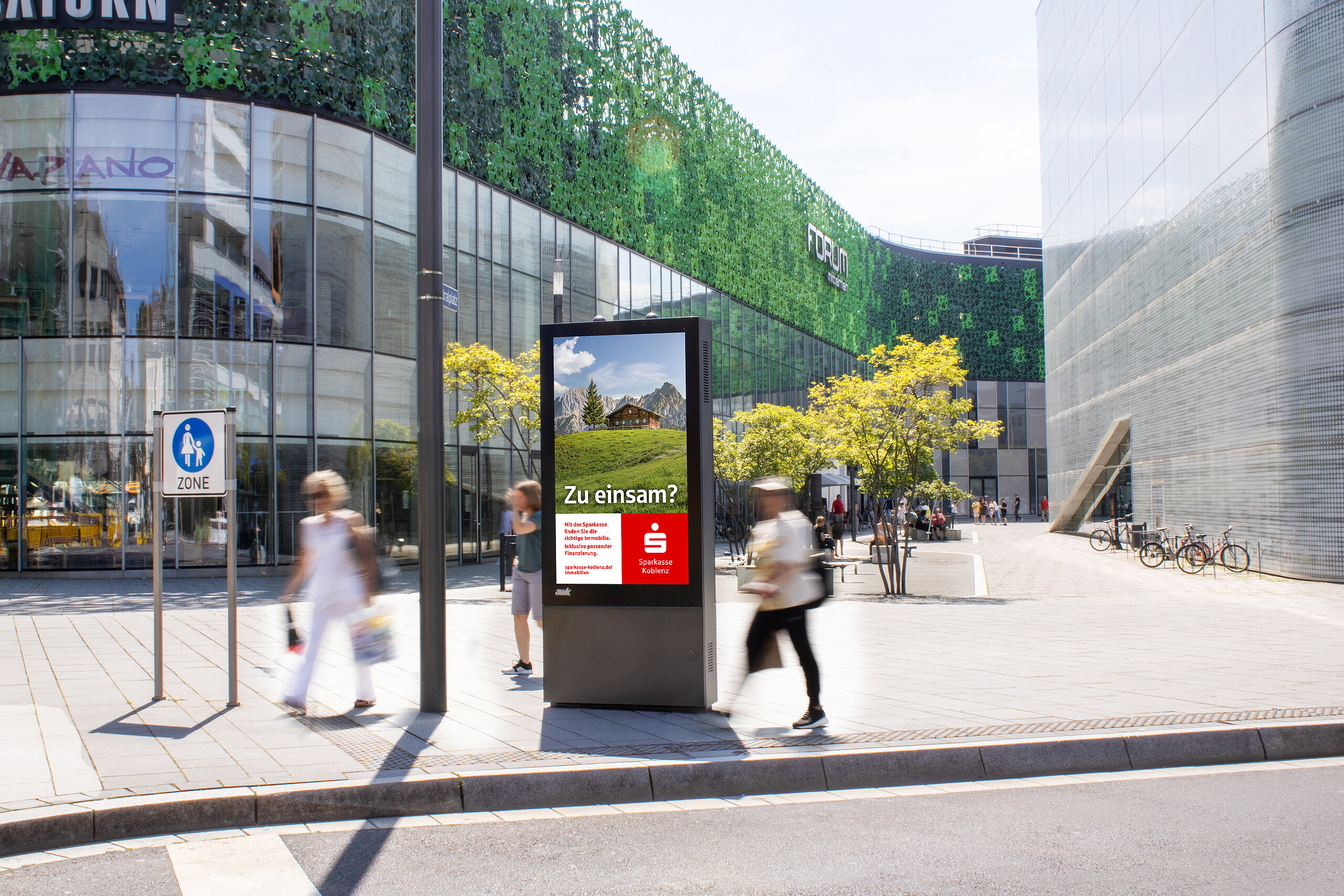 Digitale Vitrine mit Motiv der Sparkasse Koblenz vor dem Forum Koblenz, umgeben von Fußgängern in einer belebten Innenstadtzone mit moderner Architektur.