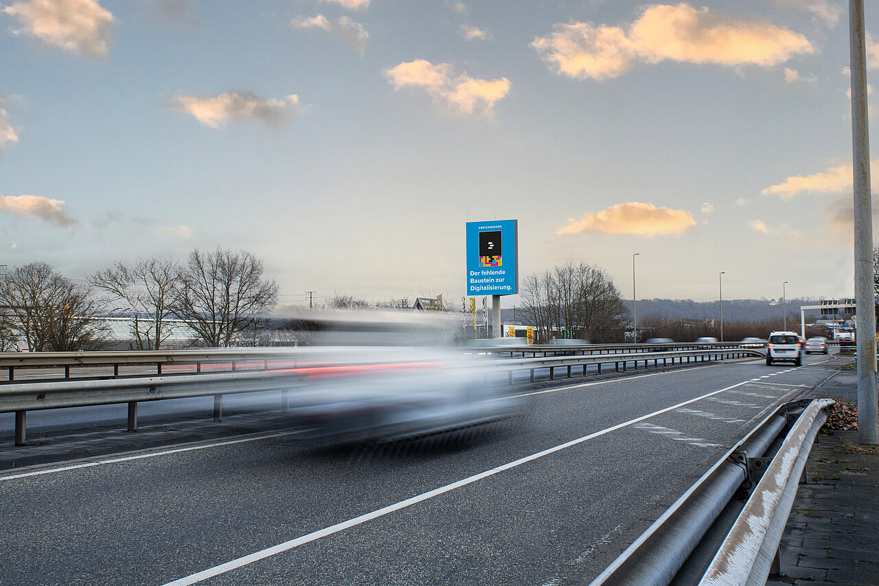 Digitaler Tower mit blauem Werbemotiv an einer stark befahrenen Straße, vorbeifahrende Autos und winterliche Umgebung im Hintergrund.