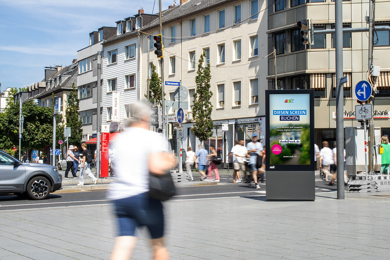 Digitale Vitrine als Werbescreen in der Koblenzer Innenstadt, umgeben von Fußgängern und Verkehr, mit Geschäften und Wohnhäusern im Hintergrund.