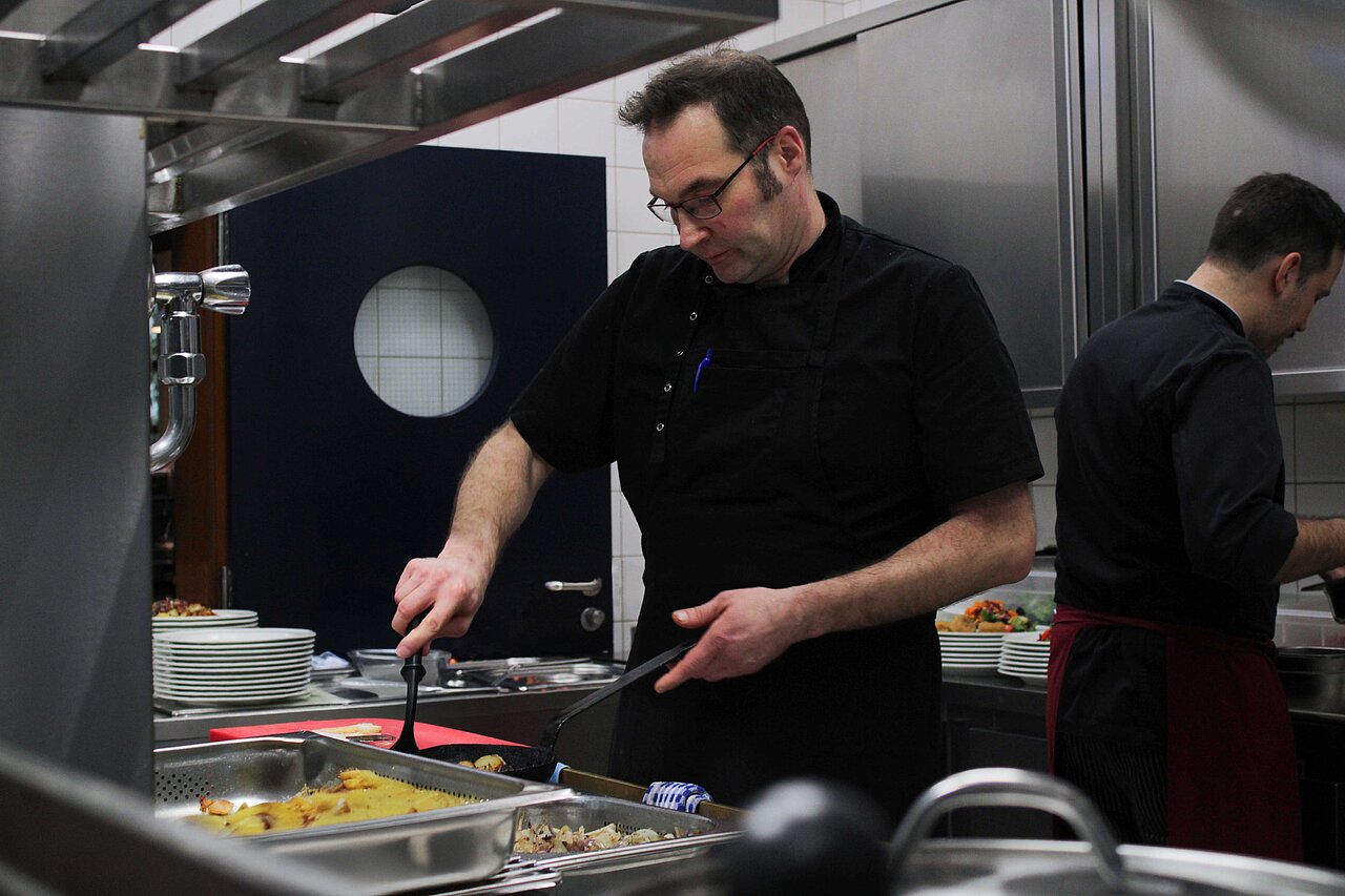 Koch bereitet in der Firmenküche frische Mahlzeiten für Mitarbeitende von Plakat-verkauft.de zu – gesunde Verpflegung als Arbeitgeber-Benefit.