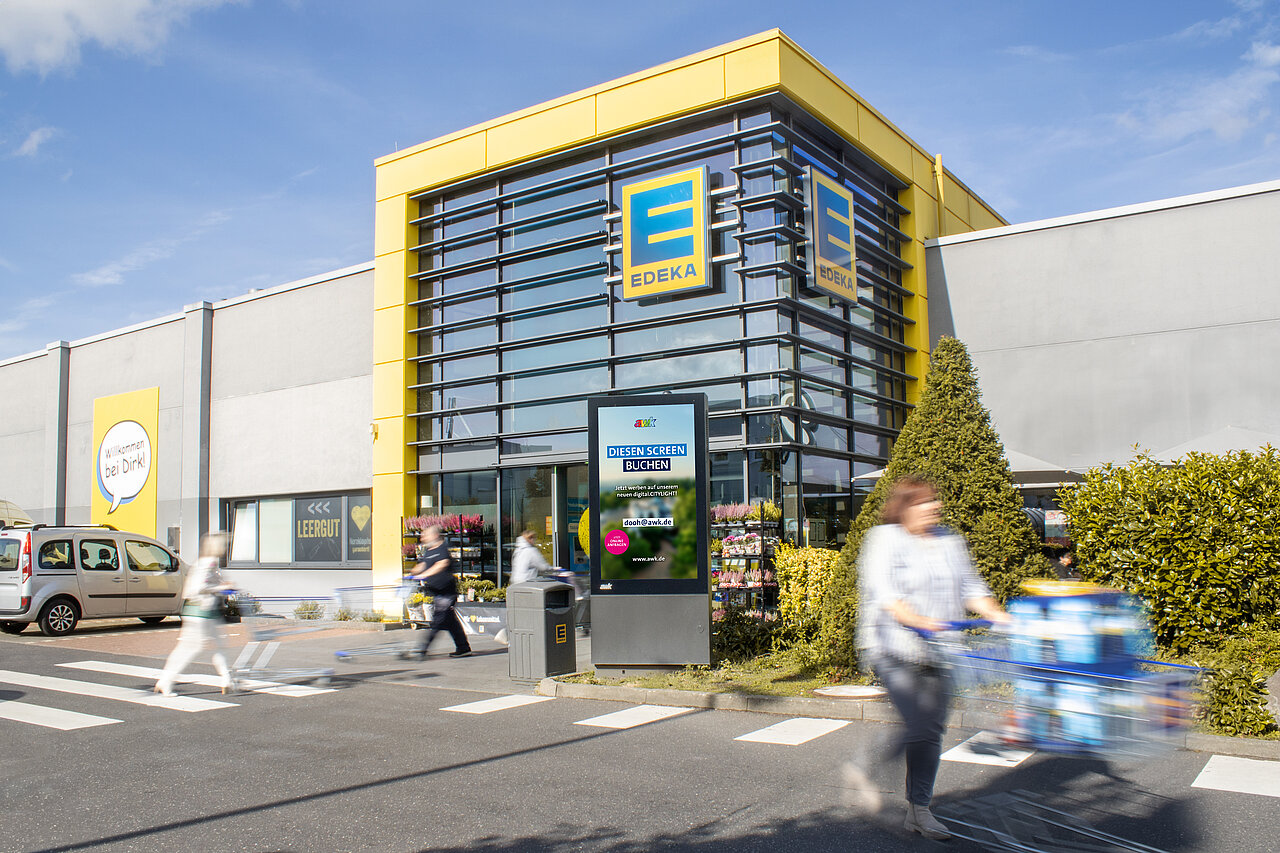Digitale Werbevitrine vor einem Supermarkt-Eingang mit vorbeilaufenden Kunden und Einkaufswagen.