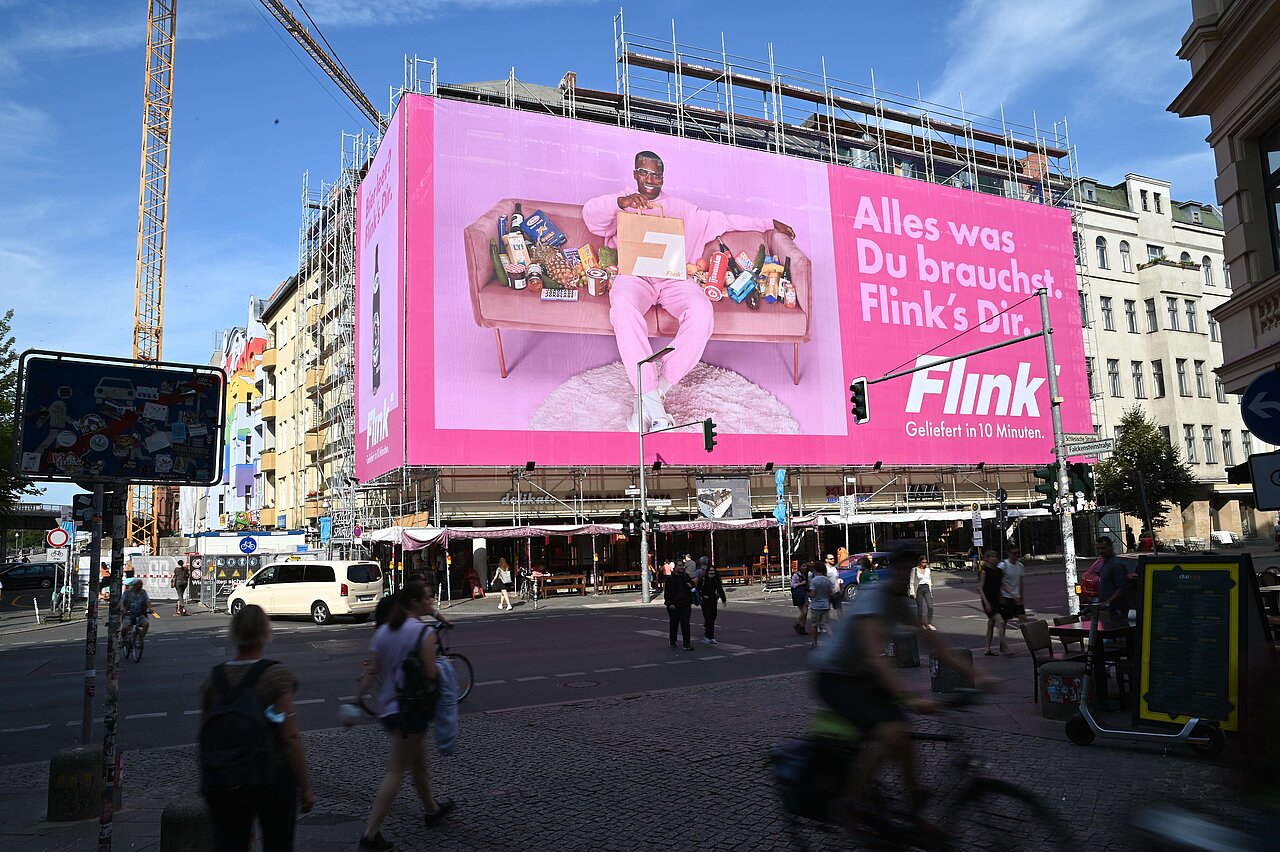 Riesenposter an einem Gebäude in der Stadtmitte, das eine auffällige Werbung für 'Flink' zeigt. Werbeplakat im XXL-Format mit einem bunten Design und einem stilvollen Foto des Modells.