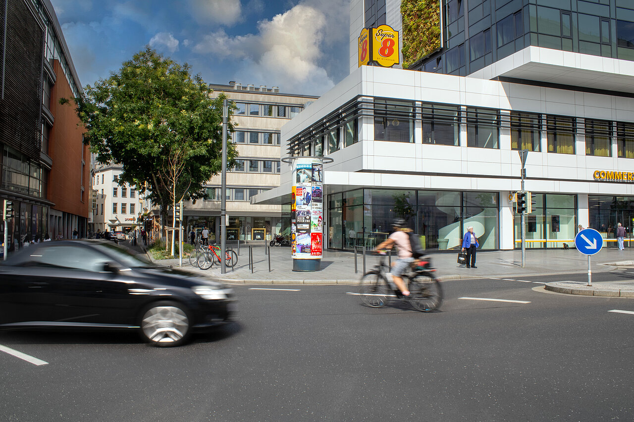 Plakatsäule im städtischen Kreuzungsbereich mit mehreren bunten Veranstaltungsplakaten, Autos und Fahrrädern vorbeifahrend