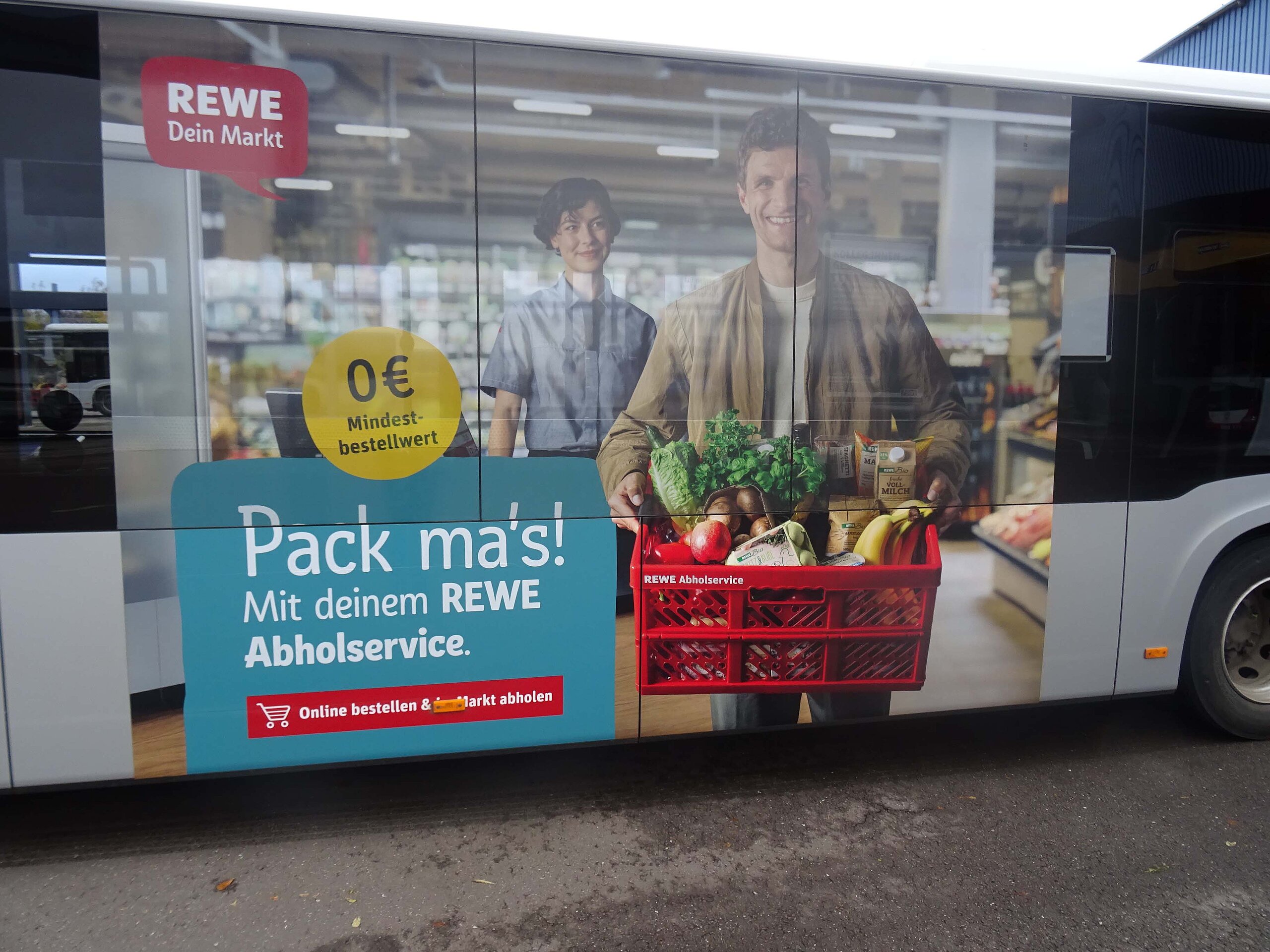 Traffic Board auf einem Bus für REWE mit dem Abholservice, zeigt einen Kunden mit einer Einkaufskiste und dem Slogan „Pack ma’s!“.