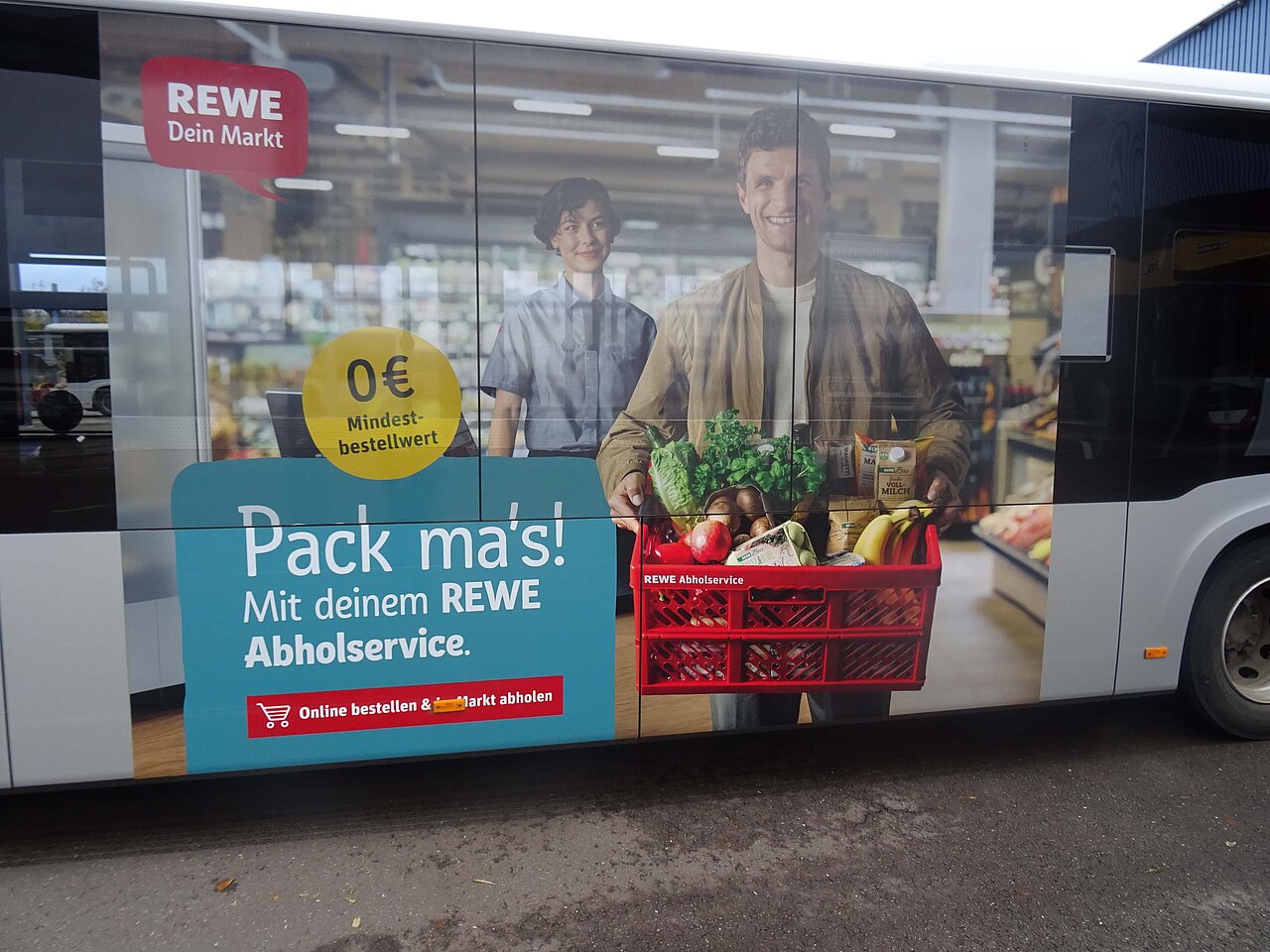 Traffic Board auf einem Bus für REWE mit dem Abholservice, zeigt einen Kunden mit einer Einkaufskiste und dem Slogan „Pack ma’s!“.