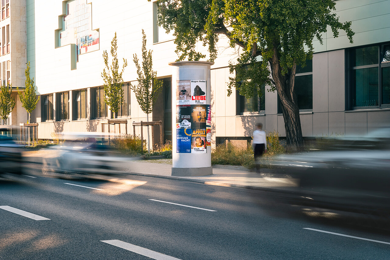 Plakatsäule an einer städtischen Straße bei Tageslicht mit mehreren Veranstaltungsplakaten