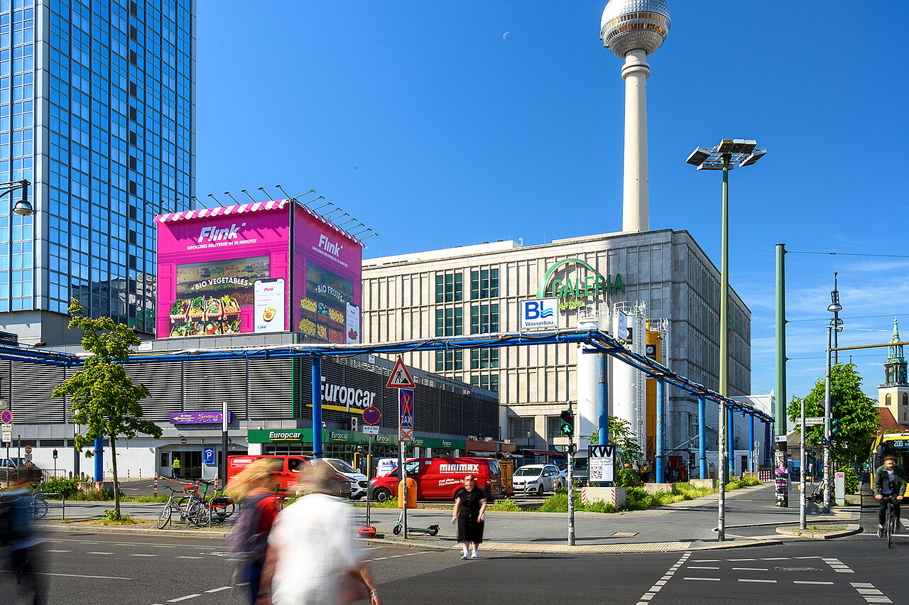 Große pinke Flink-Werbeinstallation im Stil eines Kiosks auf einem Gebäudedach am Berliner Alexanderplatz, mit Fernsehturm und belebter Kreuzung im Hintergrund.