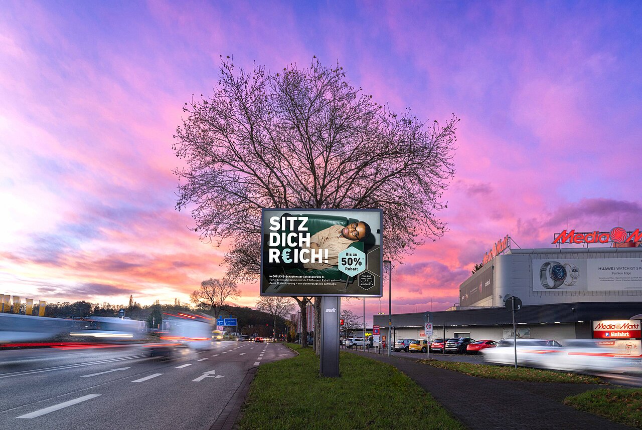 Großes Werbeplakat in einer belebten Straße mit dem Motiv eines Mannes auf einem Sofa und dem Slogan 'SITZ DICH REICH!'. Das Plakat ist an einer gut sichtbaren Stelle im Stadtverkehr angebracht und zieht die Aufmerksamkeit der vorbeifahrenden Autos und Fußgänger auf sich.