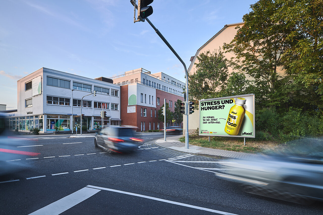 Werbeplakat für yfood mit dem Slogan 'Stress und Hunger?' und einem Bild der Produktflasche. Das Plakat ist an einer belebten Straßenecke gut sichtbar für Autofahrer und Passanten.