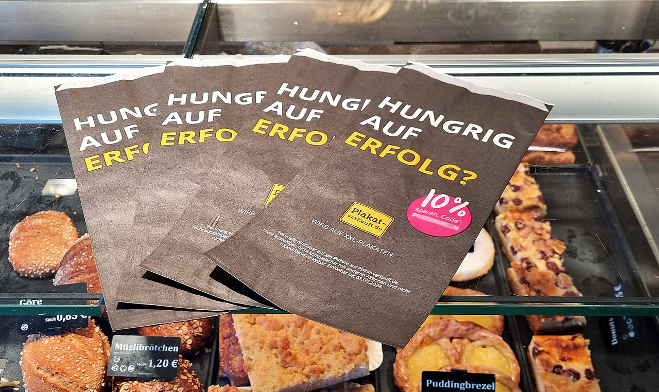 Mehrere bedruckte Brötchentüten mit „Hungrig auf Erfolg?“ liegen in einer Bäckerauslage