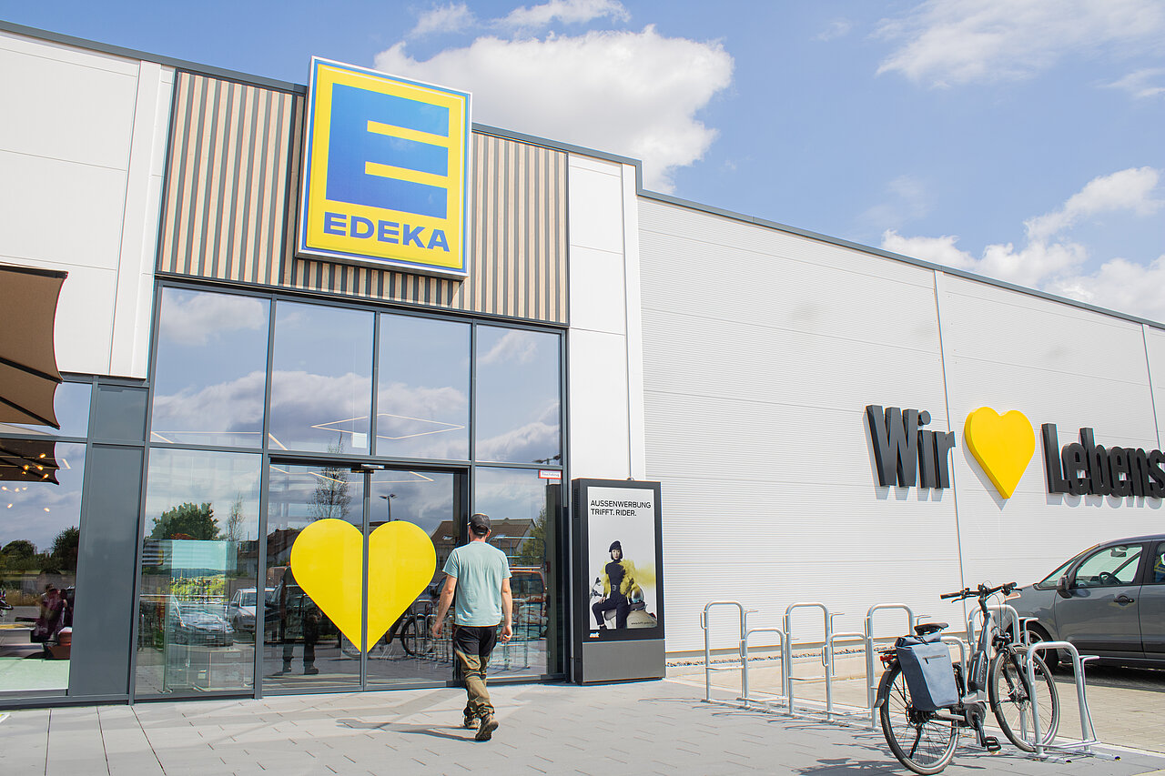 Digitale Vitrine vor einem Supermarkt-Eingang mit Werbemotiv eines sportlichen Riders, während ein Kunde auf den Eingang zuläuft.
