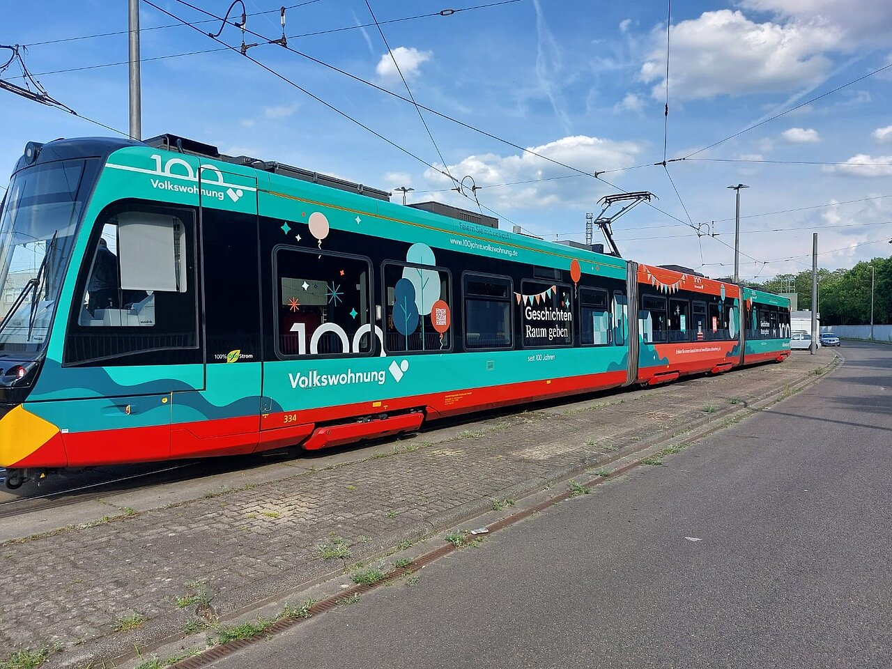 Farbenfrohe Straßenbahn im Volkswohnung-Design zum 100-jährigen Jubiläum, mit grafischen Motiven und Schriftzügen, stehend an einer Straßenbahnhaltestelle.