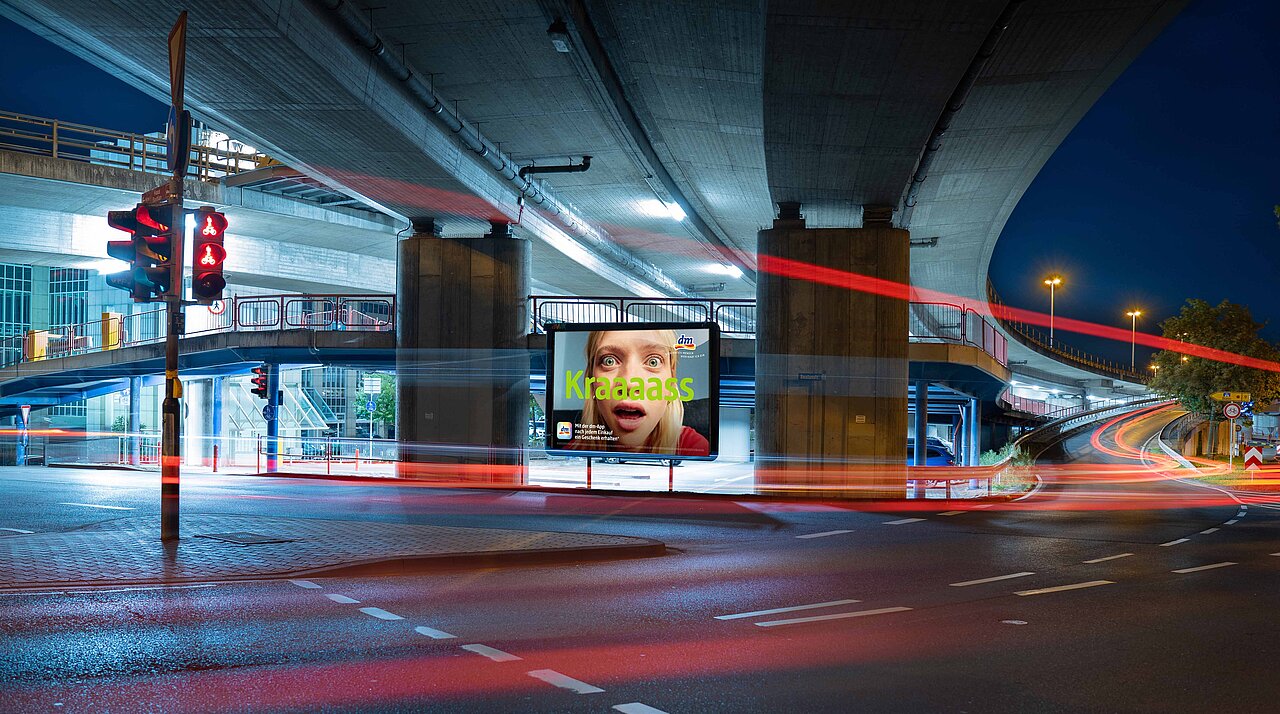 Beleuchtetes Großplakat unter einer Straßenbrücke mit auffälligem „Kraaaass“-Motiv, umgeben von vorbeiziehenden Autos und Lichtspuren – buchbar über Plakat-verkauft.de.
