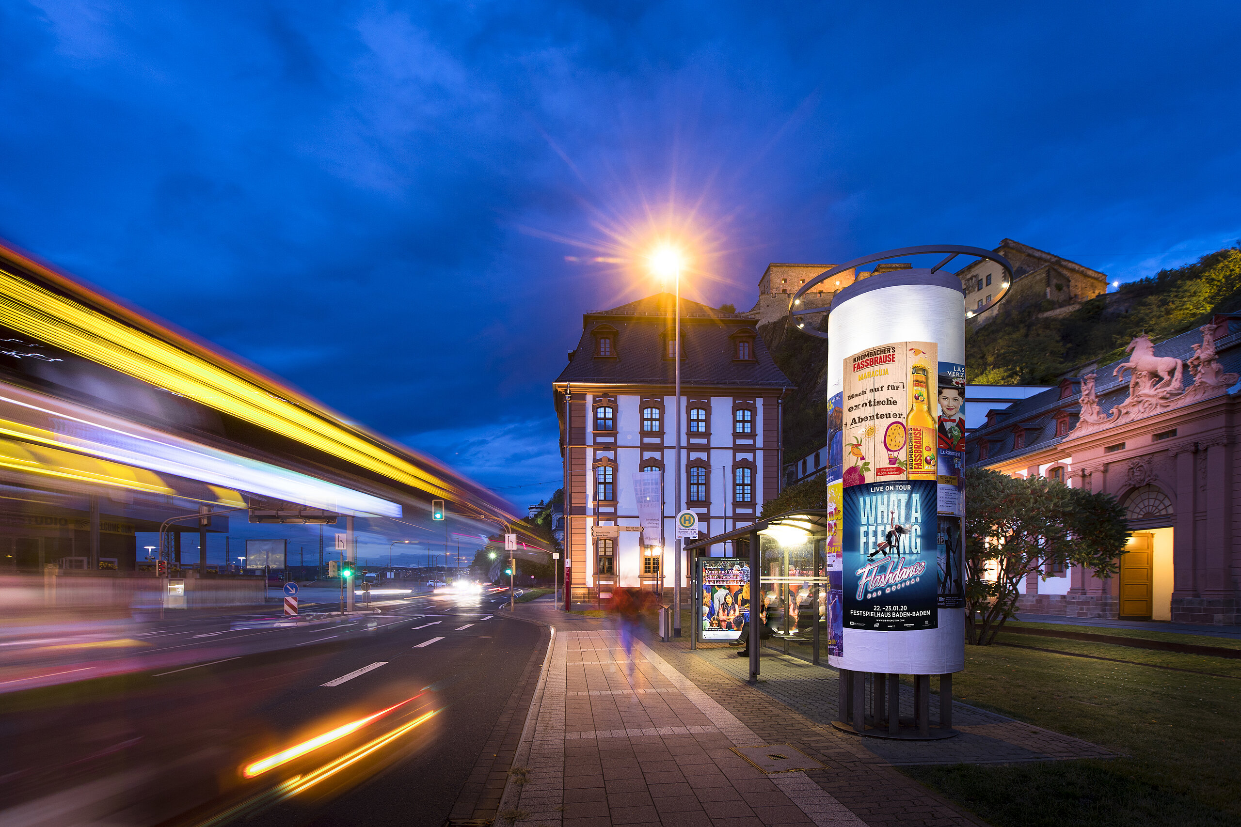 Beleuchtete Plakatsäule an einer Straße bei Nacht, mit vorbeifahrendem Bus und Stadtgebäuden im Hintergrund