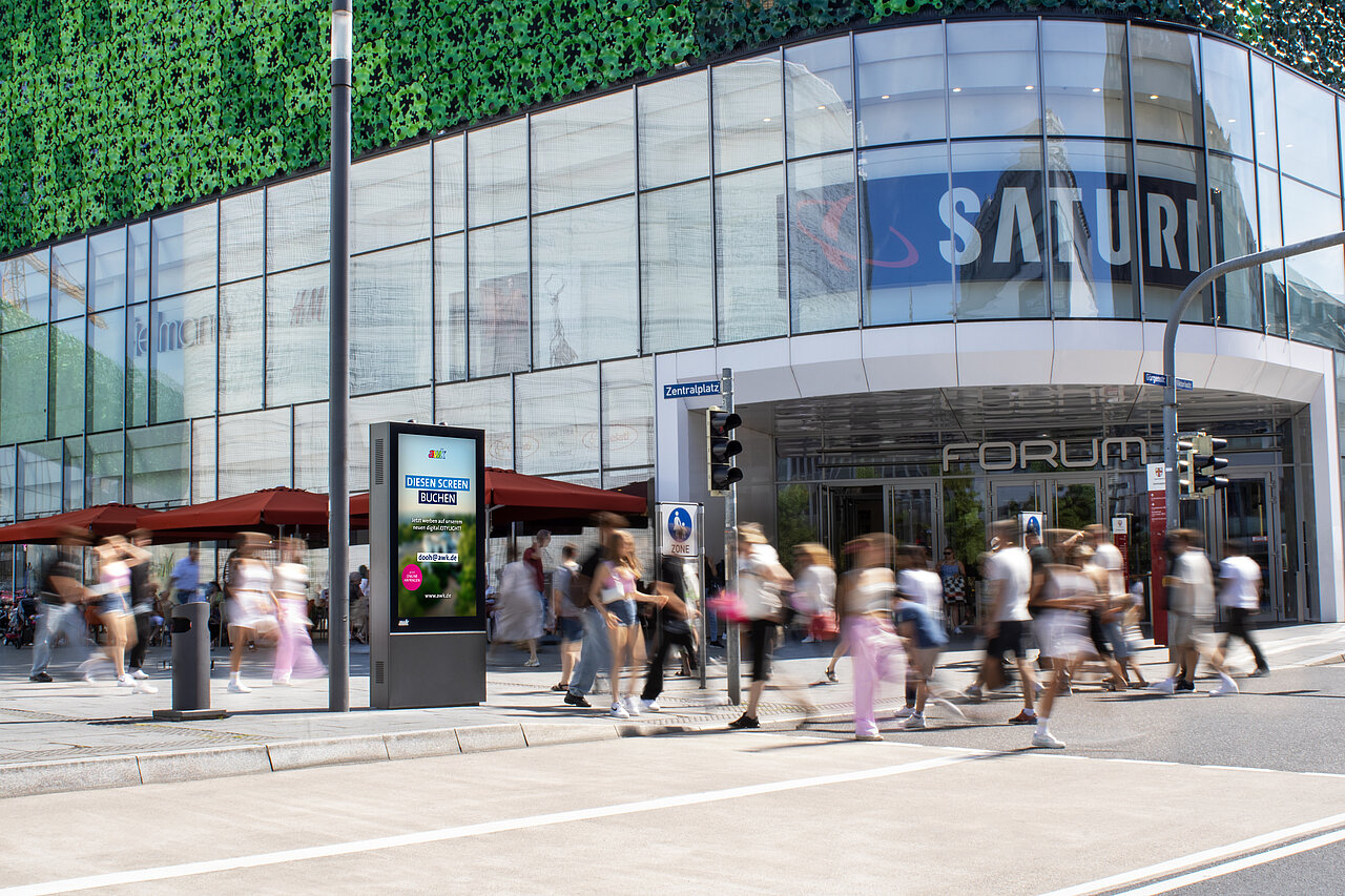 Digitale Werbevitrine vor dem Forum-Einkaufszentrum, umgeben von vielen vorbeilaufenden Passanten im Stadtzentrum.
