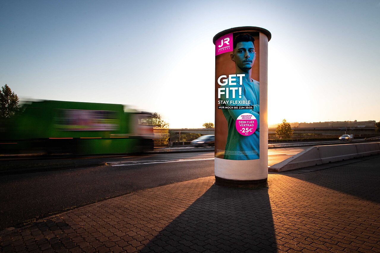 Ganzsäule mit Werbeanzeige für JR Fitness in der Abenddämmerung, umgeben von Fahrzeugen auf der Straße. Maximale Sichtbarkeit auf 360°-Fläche.