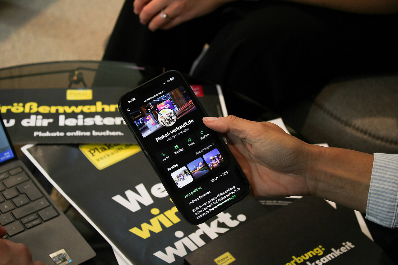 Hand hält Smartphone mit geöffnetem WhatsApp-Account von Plakat-verkauft.de – im Hintergrund Werbematerialien mit Unternehmenslogo.