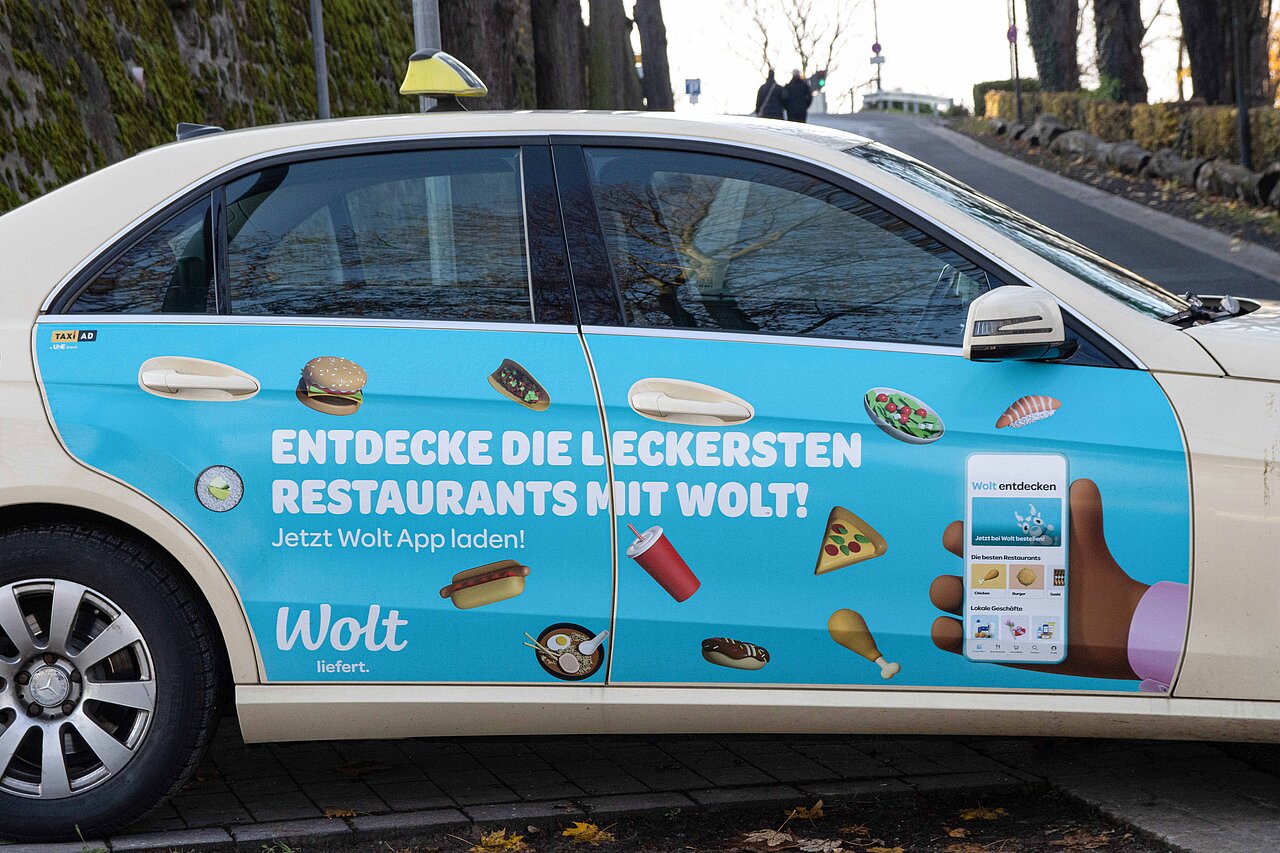 Wolt Taxiwerbung auf einem Mercedes-Benz mit Bildern von leckeren Gerichten wie Pizza und Sushi. Werbung für die Wolt App und schnelle Essensbestellungen.