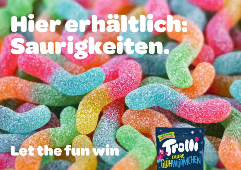 Plakatmotiv der Marke Trolli mit bunten sauren Glühwürmchen-Gummis und dem Werbespruch „Hier erhältlich: Saurigkeiten – Let the fun win“.