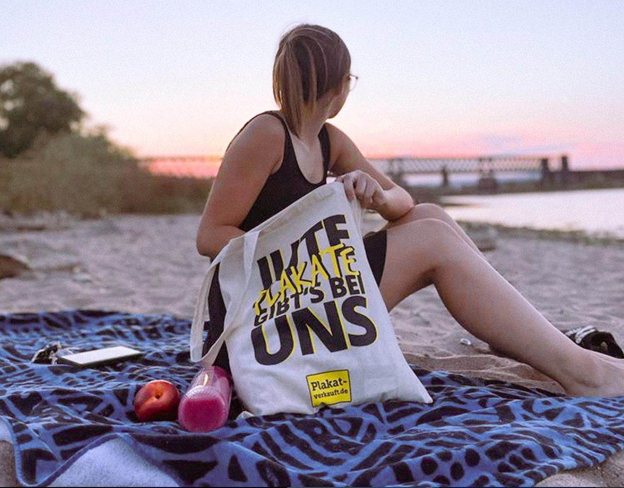 Mitarbeiterin von Plakat-verkauft.de sitzt entspannt am Strand mit einem gebrandeten Jutebeutel