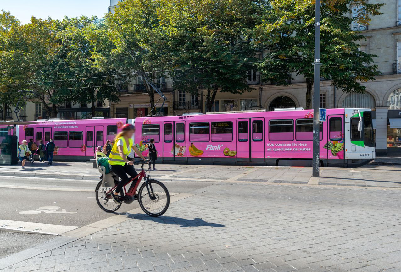 Pinke Straßenbahn mit großflächiger Flink-Werbung, gestaltet mit Obstmotiven und Slogan, unterwegs in einer belebten Innenstadtstraße mit Radfahrerin im Vordergrund.