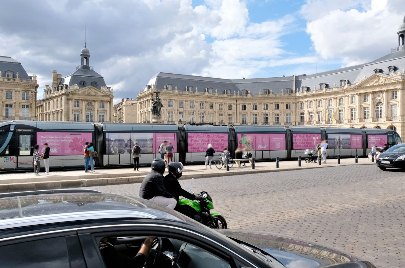 Straßenbahn mit pinker Flink-Vollbeklebung vor eindrucksvollen historischen Gebäuden, umgeben von Passanten, Radfahrern und Verkehr im urbanen Umfeld.