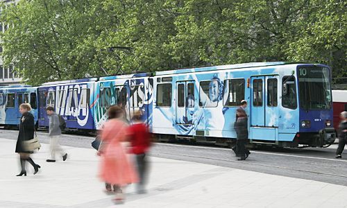Straßenbahn in blauem VILSA-Design mit Wasser- und Markenmotiven, vorbeigehende Passanten in Bewegung, Bäume im Hintergrund.