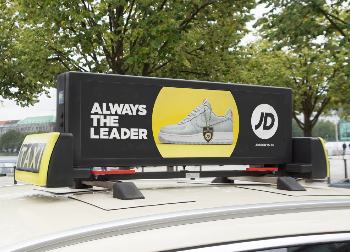 Taxi-Dachwerbung mit dem Slogan 'ALWAYS THE LEADER' und einem Bild von weißen Sneakern von JD Sports. Perfekte Werbung auf dem Dach eines Taxis, das durch die Stadt fährt.