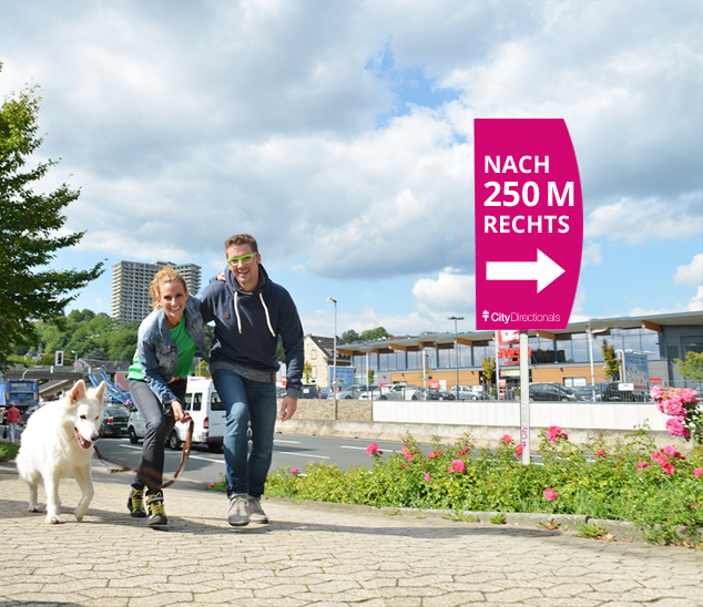 Werbeschild mit Richtungshinweis in einer belebten Gegend. Ein Werbeschild zeigt den Weg nach rechts, während eine Familie mit Hund spaziert.